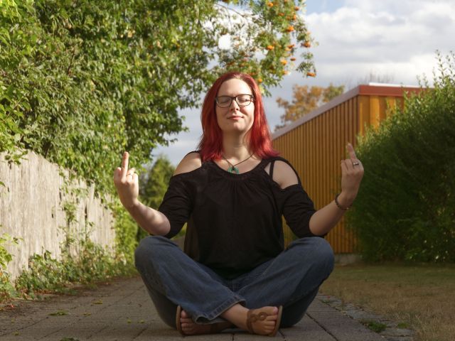 Elke Sommer, die im Schneidersitz in einer friedlichen Umgebung im Freien sitzt und eine meditative Haltung mit einem spielerischen Ausdruck einnimmt.